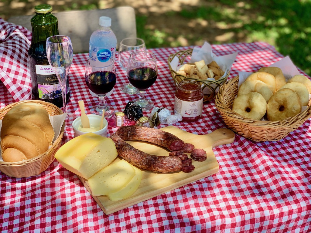 mesa de piquenique, com queijo, salame, pão, suco de uva, roda de carreta e geleias