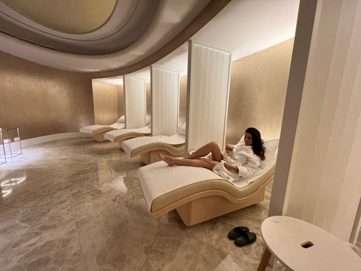 Na foto a jornalista Ane Zanoni, uma mulher branca de cabelos pretos, relaxa no spa do hotel four seasons, deitada em uma das espreguiçadeiras que estão dispostas no local. O ambiente tem cores suaves, em tons nudes, com luz baixa e muito aconchego