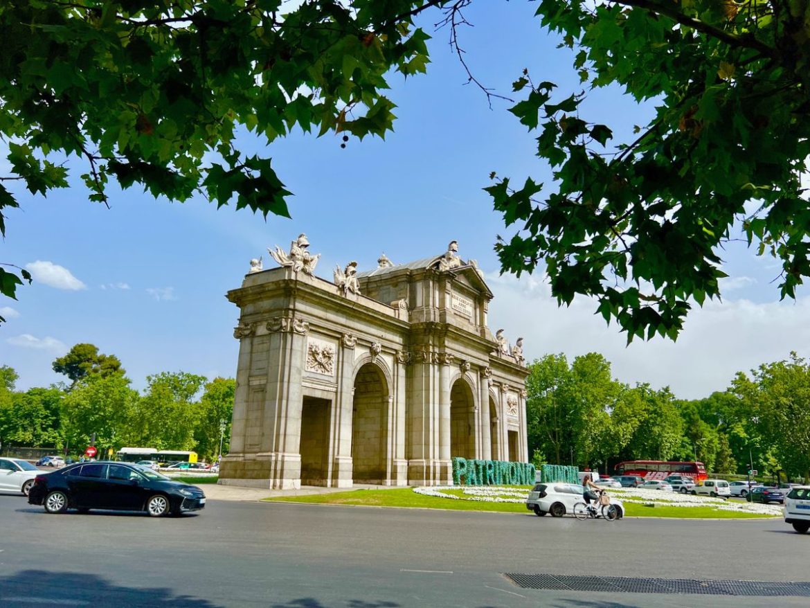 Uma grande porta construída com pedras é o foco principal da fotografia. O monumento se chama Puerta de Alcalá e fica em Madri
